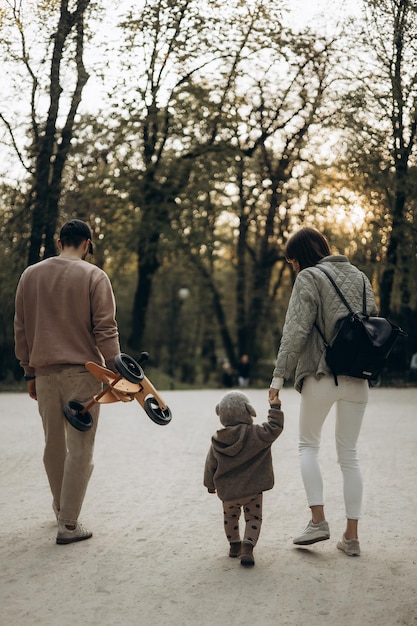 Uma foto da parte de trás de uma jovem família onde uma esposa pai e seu filho pequeno estão caminhando no parque com uma bicicleta infantil nas mãos