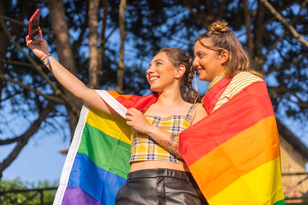 Uma foto de close de duas jovens caucasianas se abraçando com a bandeira do orgulho LGBT tirando uma selfie ao ar livre