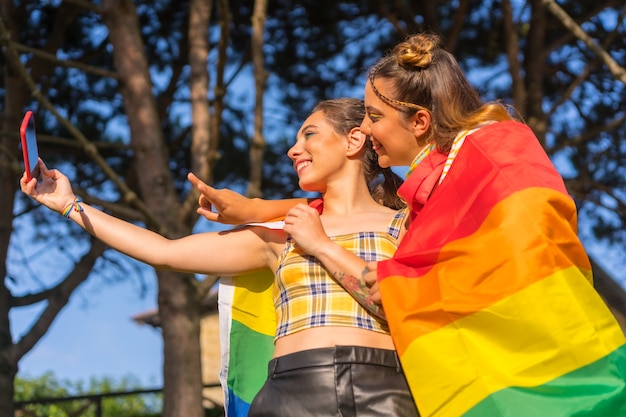 Uma foto de close de duas jovens caucasianas se abraçando com a bandeira do orgulho LGBT tirando uma selfie ao ar livre