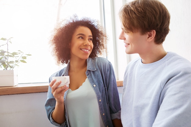 Foto uma foto de dois estudantes sentados juntos muito perto. menina está segurando um copo nas mãos e olhando para o cara. ele está olhando para ela e sorrindo.