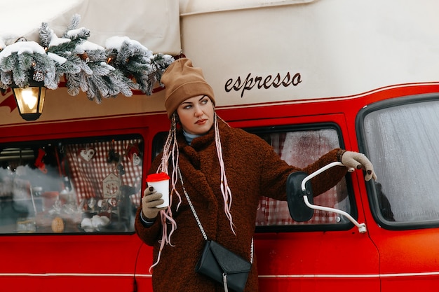 Uma foto de rua de uma jovem mulher com um casaco e boné uma xícara de café na mão e olhando para longe.