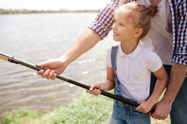 Uma foto do cara ajudando a filha a segurar a vara de peixe da maneira certa. Menina está segurando com as duas mãos e sorrindo. Ela parece feliz.