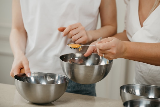 Uma foto próxima das mãos de duas mulheres, uma delas movendo a gema em uma colher para outra tigela de sopa de aço inoxidável na cozinha.