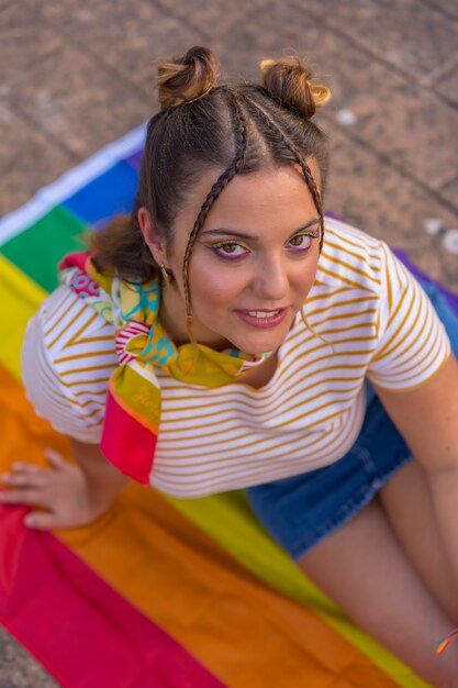 Uma foto vertical de uma jovem mulher branca sentada na bandeira do orgulho LGBT ao ar livre