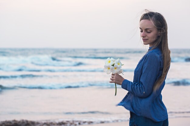 Uma garota à beira-mar ao pôr do sol, segurando uma flor de plumeria na mão