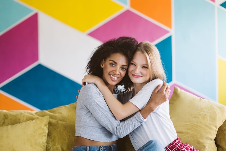Foto uma garota afro-americana bonita com cabelo escuro encaracolado e uma garota sorridente com cabelos loiros se abraçando alegremente enquanto alegremente com uma parede colorida