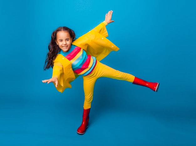 Uma garota alegre com uma capa de chuva amarela e botas de borracha vermelhas pula com os braços estendidos e olha para a câmera.