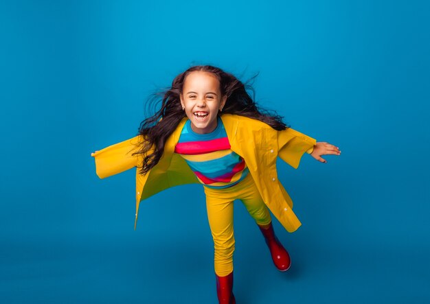 Uma garota alegre com uma capa de chuva amarela e botas de borracha vermelhas pula com os braços estendidos e olha para a câmera.