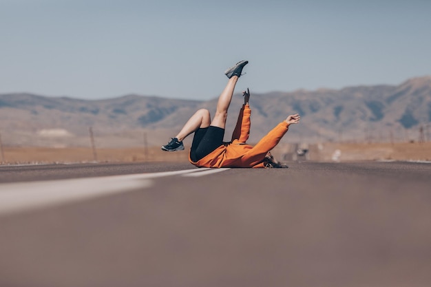 Uma garota alegre em um capuz laranja posa brilhantemente deitada na estrada contra o pano de fundo de grandes montanhas rochosas altas