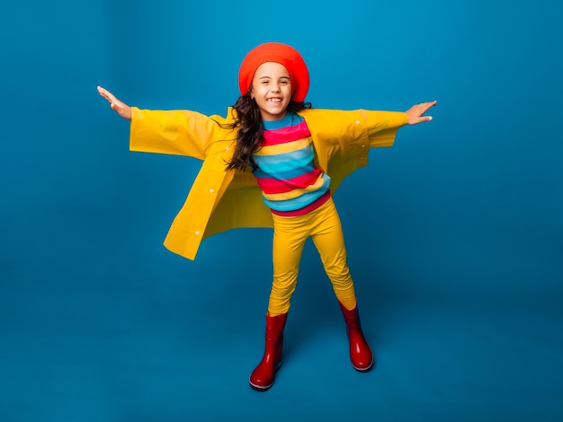 Uma garota alegre em uma capa de chuva amarela, boina vermelha e botas de borracha pula com os braços estendidos e olha para a câmera.