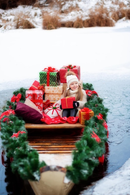 Uma garota anda em um lago em um barco de Natal Perto há uma floresta de inverno com neve e muitos presentes de