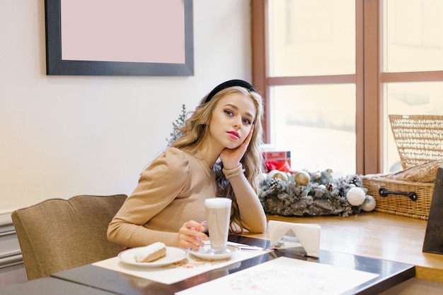 Uma garota atraente e feliz está sentada em um café decorado para o Natal, bebendo café e comendo sobremesa