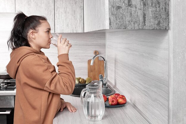 Uma garota bebe água limpa de um copo em casa na cozinha