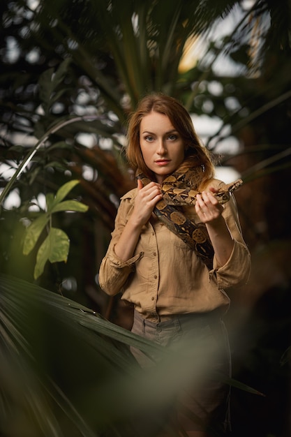 Uma garota bonita com maquiagem natural e cabelo vermelho fica na selva entre plantas exóticas com cobra.