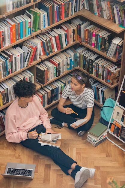 Uma garota bonita e seu colega de classe sentados no chão em uma grande estante na biblioteca da faculdade enquanto discutem o enredo de um romance