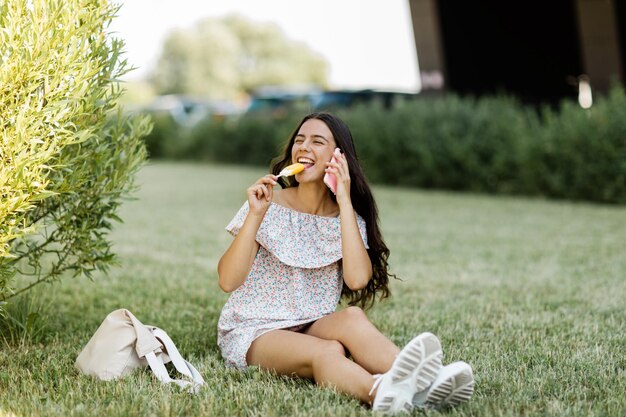 Foto uma garota bonita está falando no telefone sentada na grama e comendo sorvete