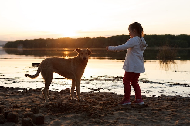 Uma garota brinca com um cachorro em uma praia à beira do rio ao pôr do sol.