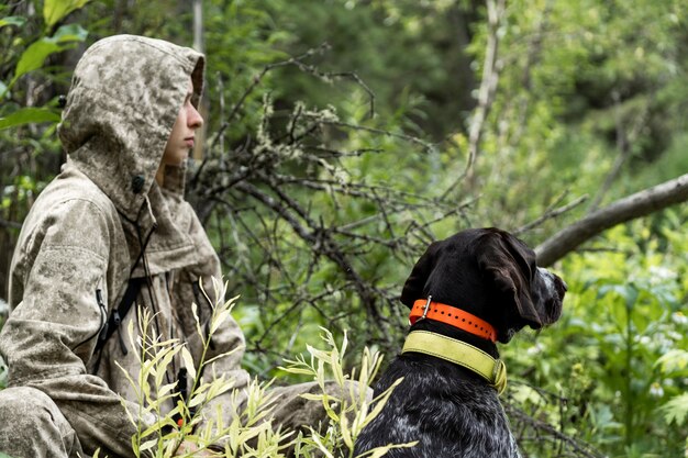 Uma garota caçadora senta-se com seu cachorro nos arbustos. Uma garota caça com um drathaar alemão