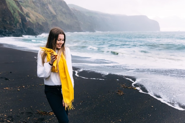 Uma garota caminha ao longo de uma praia vulcânica na Islândia Ela está vestindo calças pretas e um suéter branco com um lenço amarelo