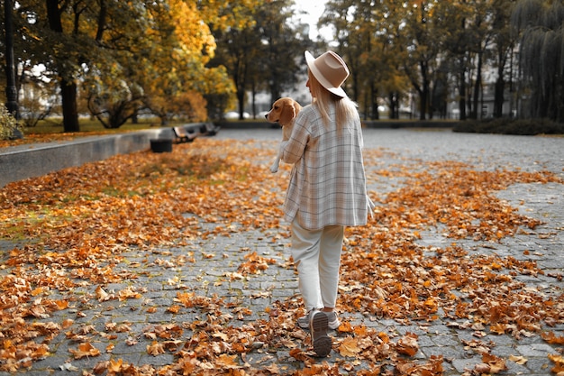 Uma garota caminha pelas folhas no outono com um cachorro nos braços.