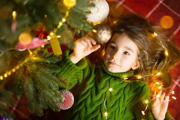 Uma garota com cabelo comprido e guirlandas encontra-se em uma manta vermelha sob uma árvore de natal com brinquedos em um agasalho de malha quente. natal, reveillon, emoções infantis, alegria, expectativa de um milagre e presentes