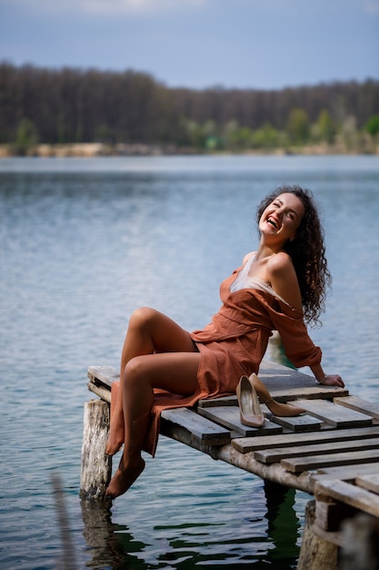 Uma garota com cabelo encaracolado longo ondulado em um vestido longo guipura com os pés descalços no verão em uma floresta em um lago ao pôr do sol em pé sobre um pantone em uma ponte de cais de madeira. Dia ensolarado de verão