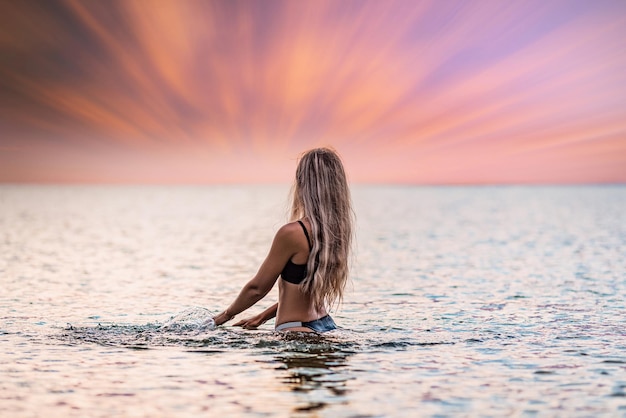 Uma garota com cabelo loiro em um maiô preto espirra para os lados enquanto está sentado em um estuário em um fundo por do sol