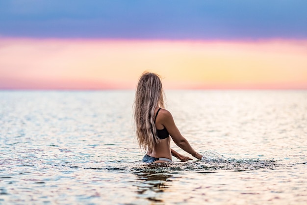 Uma garota com cabelo loiro em um maiô preto espirra para os lados enquanto está sentado em um estuário em um fundo por do sol