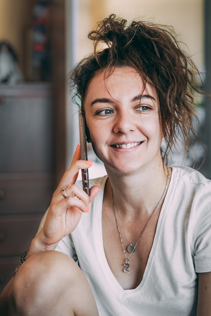 Uma garota com cabelos cacheados e camiseta branca está sentada em casa conversando ao telefone com os amigos.