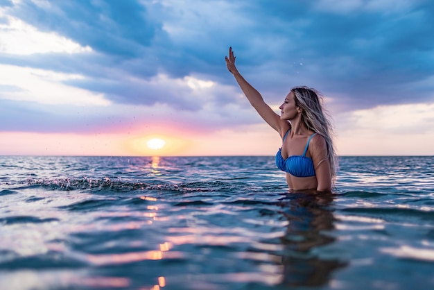 Uma garota com cabelos loiros em um maiô azul espirra para os lados enquanto está sentado em um estuário em um fundo por do sol