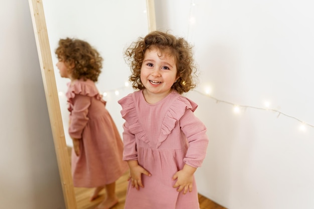 uma garota com cachos em um vestido rosa ri do espelho