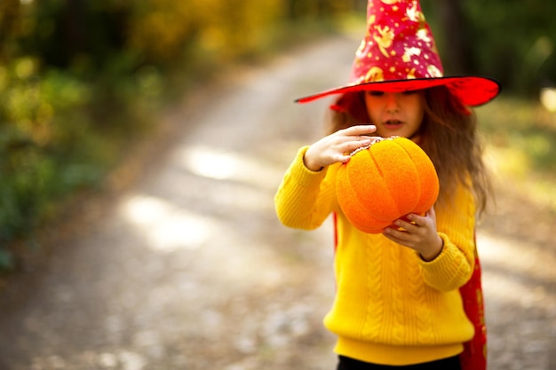 Uma garota com chapéu de bruxa em um suéter amarelo e com uma abóbora com olhos esculpidos e uma boca Jack o Lantern em uma floresta amarela de outono Halloween Holiday