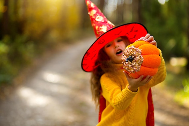 Uma garota com chapéu de bruxa em um suéter amarelo e com uma abóbora com olhos esculpidos e uma boca jack o lantern em uma floresta amarela de outono halloween holiday