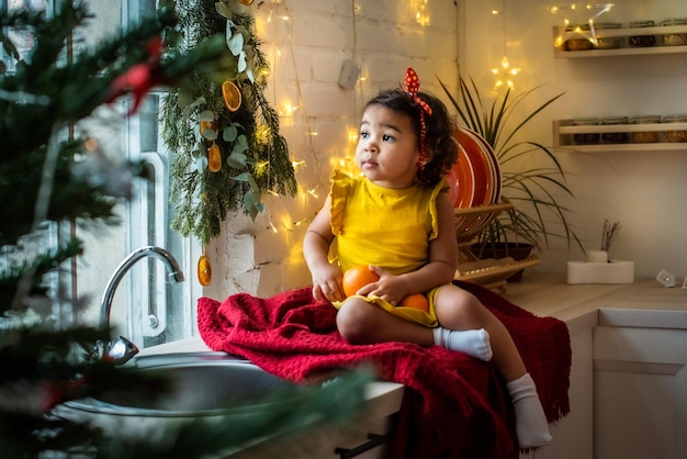 Uma garota com cor de pele escura, ela está sentada na mesa usando um vestido amarelo e segurando laranjas