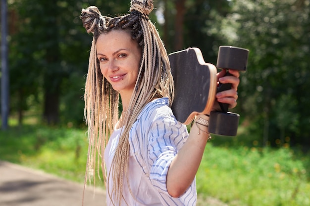 Uma garota com dreadlocks em uma camisa listrada segura um longboard nos ombros, sorri e olha diretamente para a câmera esporte moderno foto de alta qualidade