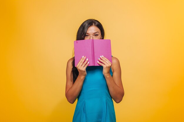 Uma garota com óculos em um fundo amarelo em um vestido azul Cobriu o rosto com um caderno rosa