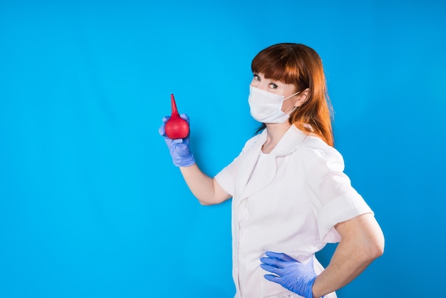 Uma garota com roupas de médico e uma máscara segura uma pêra médica nas mãos e olha para a câmera, isolada em um fundo azul