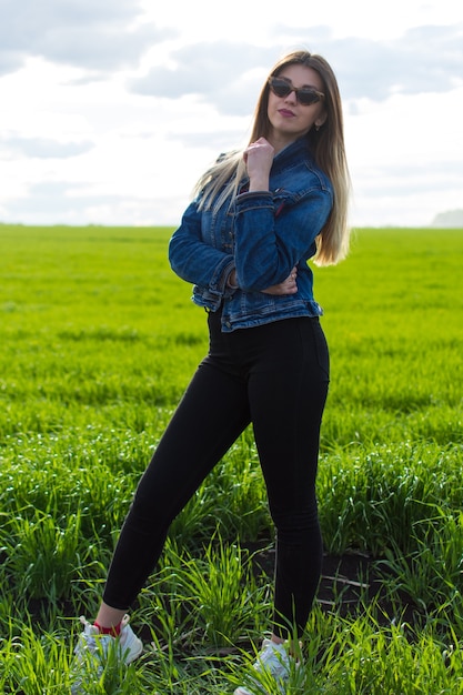 Uma garota com roupas jeans com cabelo bonito fica em um campo verde com um olhar sonhador no rosto.