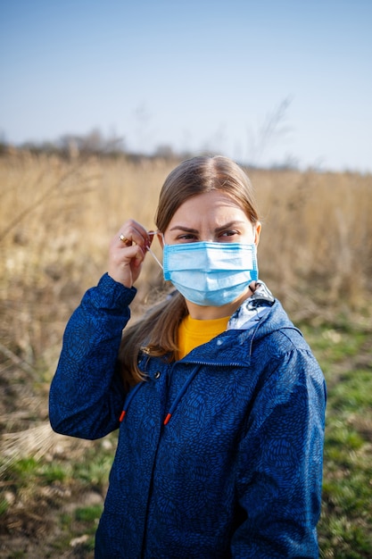 Uma garota com um casaco azul na natureza coloca uma máscara médica protetora. Máscara de dia ensolarado ao ar livre no rosto contra vírus