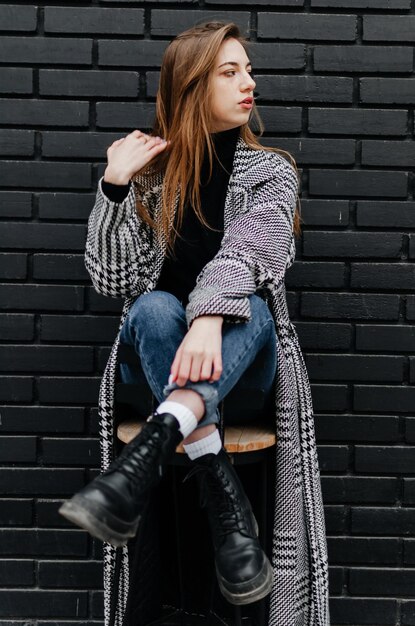 uma garota com um casaco cinza senta-se em uma cadeira perto de uma parede preta