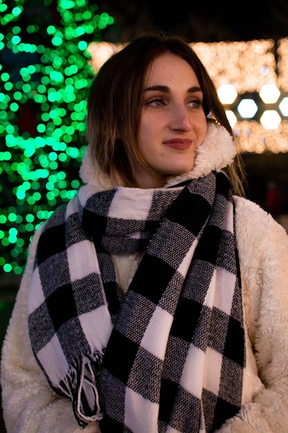 Uma garota com um casaco e luvas está embaixo de uma árvore de Natal decorada com luzes. Ano Novo de 2022.