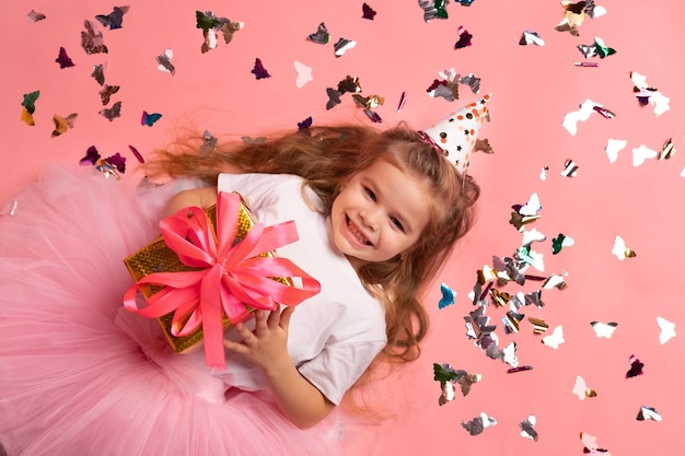 Uma garota com um chapéu de aniversário segura um presente na frente de um fundo rosa com borboletas.