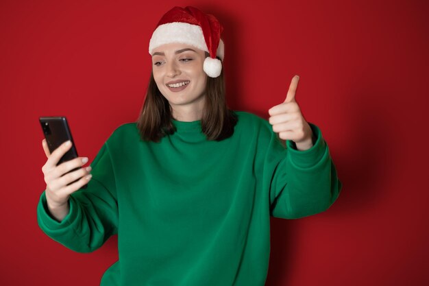 Uma garota com um chapéu de ano novo está falando ao telefone em um fundo liso segurando um telefone na mão pedindo jantar o conceito de fim de semana de ano novo 2023 feliz natal 2023 feriados