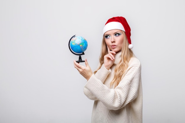 Uma garota com um chapéu de Papai Noel com um globo pensa sobre um fundo cinza