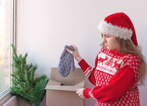 Uma garota com um chapéu de papai noel e um suéter de natal coloca luvas em uma caixa ao lado de uma pequena árvore de natal