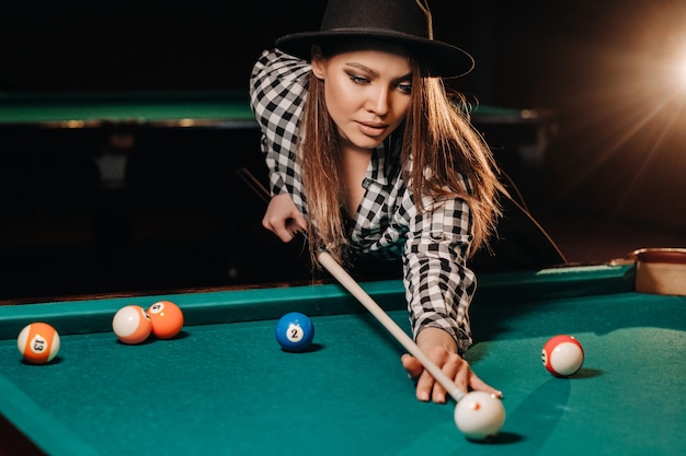 Uma garota com um chapéu em um clube de bilhar com um taco nas mãos acerta uma bola. Jogando sinuca.