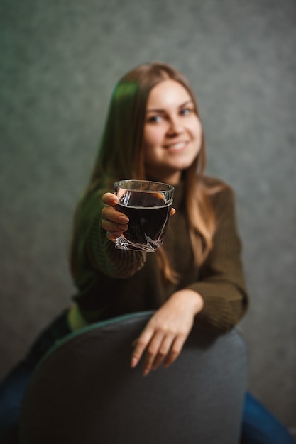 Uma garota com um copo com um líquido preto sentada em uma cadeira em um fundo cinza