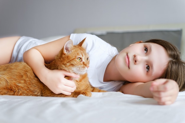 Uma garota com um gato ruivo deita-se em uma cama branca. Uma menina acariciando um gato ruivo enquanto está deitada em um lençol branco