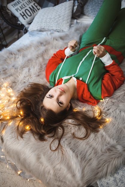 Uma garota com um macacão de ano novo e um clima festivo deitada em uma cama ao redor de guirlandas luminosas