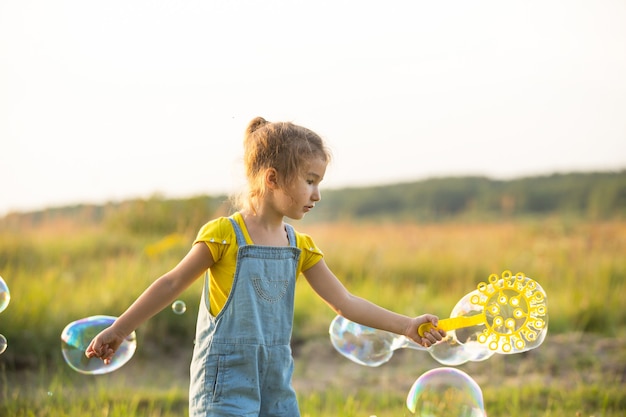 Uma garota com um macacão jeans sopra bolhas de sabão no verão em um campo ao pôr do sol. Dia Internacional da Criança, criança feliz, atividades ao ar livre. Fundo de verão. Estilo de vida saudável e ecológico